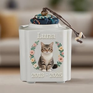 Puede incluir: Una urna blanca de cerámica para mascotas con un diseño de corona floral. La urna presenta una ilustración de un gato atigrado sentado, el nombre "Luna" y los años "2013-2025". Tiene una tapa decorativa con una tela estampada y un cordón marrón.