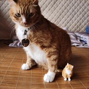 K&ouml;nnte beinhalten: Eine gro&szlig;e orange Tabby-Katze mit wei&szlig;en Abzeichen sitzt auf einer braunen geflochtenen Matte. Eine kleinere orange Tabby-Katzenfigur sitzt vor der gr&ouml;&szlig;eren Katze.