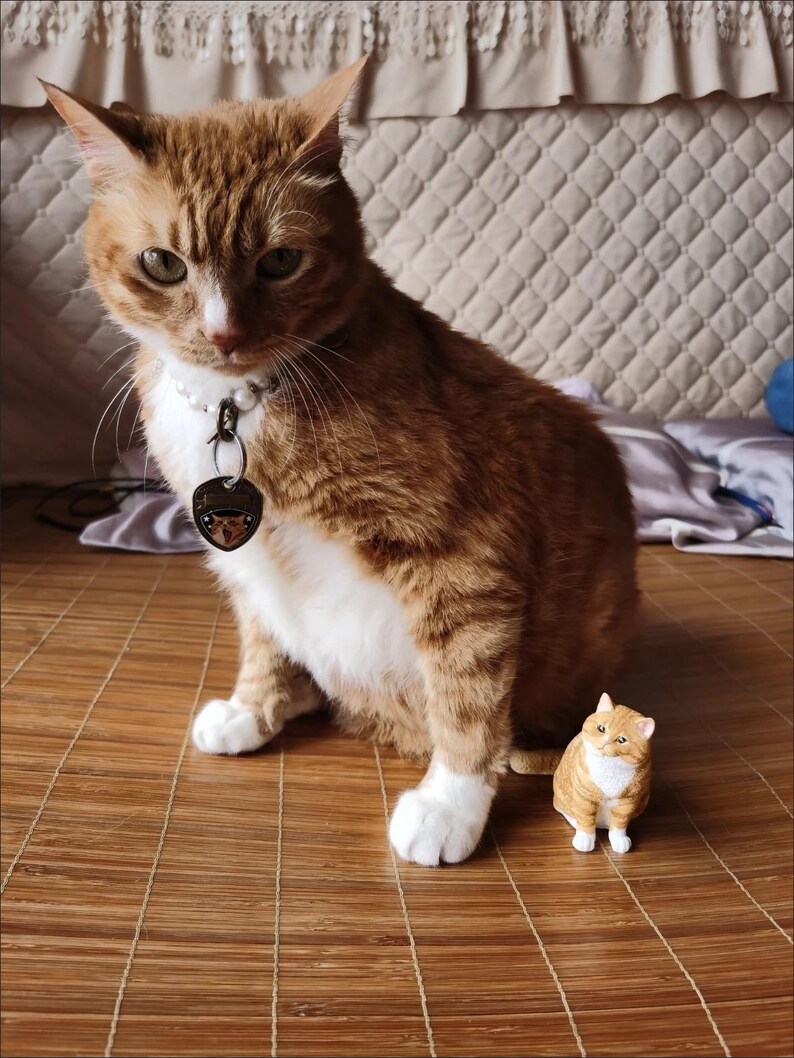 K&ouml;nnte beinhalten: Eine gro&szlig;e orange Tabby-Katze mit wei&szlig;en Abzeichen sitzt auf einer braunen geflochtenen Matte. Eine kleinere orange Tabby-Katzenfigur sitzt vor der gr&ouml;&szlig;eren Katze.