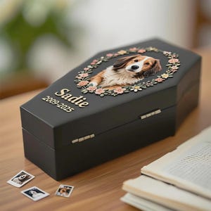 May include: A black pet memorial box shaped like a coffin, featuring a portrait of a dog surrounded by a floral wreath. The name "Sadie" and the years "2009-2025" are inscribed on the box. Small photos of the dog are scattered nearby.