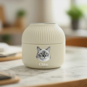 以下が含まれることがあります： 猫の顔のグレースケール画像が特徴のクリーム色のペット用骨壺。骨壺はリブ編みのトップで、猫の名前「Chloe」が画像の下に2015年から2023年までの年号とともに印刷されています。