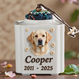 May include: A white ceramic pet urn with a 3D image of a golden retriever, surrounded by small daisy accents. The name "Cooper" and the years "2011 - 2025" are displayed below the dog's image. The lid is topped with a patterned fabric and a wooden bead.