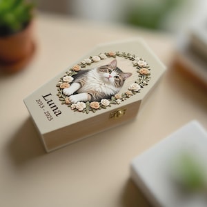 May include: A beige wooden pet memorial box with a cat illustration and floral wreath. The box is coffin-shaped and has the name "Luna" and the years "2013-2025" printed on it. A gold-colored clasp secures the box.