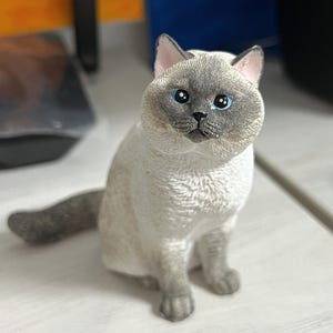 K&ouml;nnte beinhalten: Eine wei&szlig;e und graue Katzenfigur mit blauen Augen, die auf einer wei&szlig;en Oberfl&auml;che sitzt.