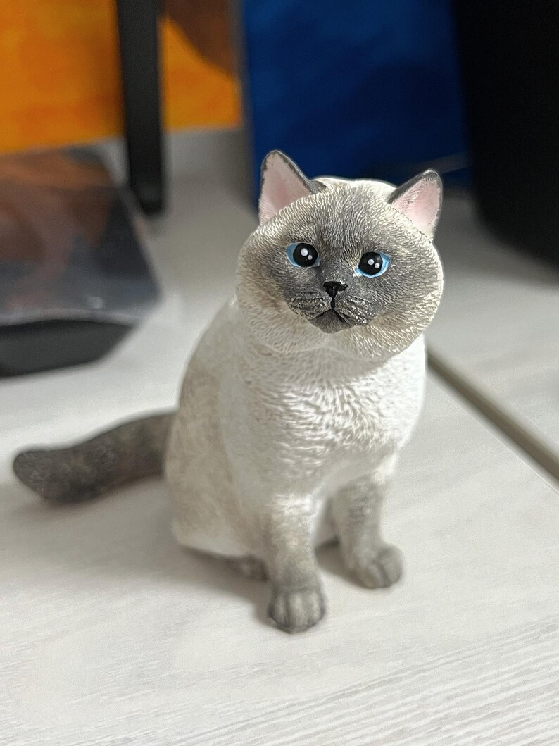 K&ouml;nnte beinhalten: Eine wei&szlig;e und graue Katzenfigur mit blauen Augen, die auf einer wei&szlig;en Oberfl&auml;che sitzt.