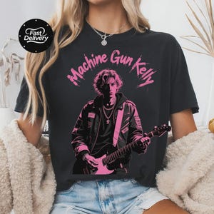Könnte beinhalten: Schwarzes T-Shirt mit einer rosa Grafik von Machine Gun Kelly, der Gitarre spielt. Der Text "Machine Gun Kelly" befindet sich über dem Bild. Ein "Schnelle Lieferung"-Logo befindet sich in der oberen linken Ecke.