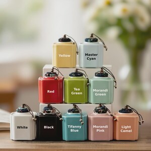 May include: A collection of ten colorful ceramic tea canisters. Each canister is square with a rounded top and a decorative lid. Colors include yellow, light blue, red, green, white, black, Tiffany blue, pink, and light camel. Text labels identify each color.