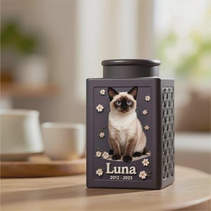Peut inclure: Urne fun&eacute;raire grise fonc&eacute;e pour animaux de compagnie, avec une illustration de chat siamois, des fleurs blanches et le nom "Luna" avec les dates 2012-2025. L'urne a un motif tiss&eacute; d&eacute;coratif et un couvercle gris fonc&eacute;.