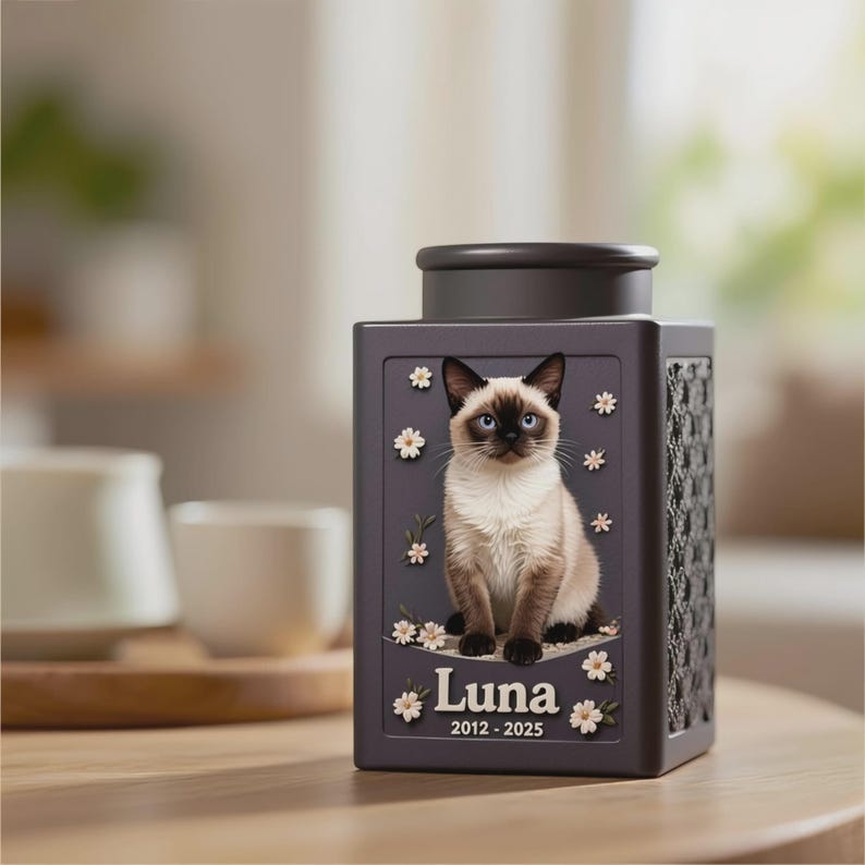 Peut inclure: Urne fun&eacute;raire grise fonc&eacute;e pour animaux de compagnie, avec une illustration de chat siamois, des fleurs blanches et le nom "Luna" avec les dates 2012-2025. L'urne a un motif tiss&eacute; d&eacute;coratif et un couvercle gris fonc&eacute;.