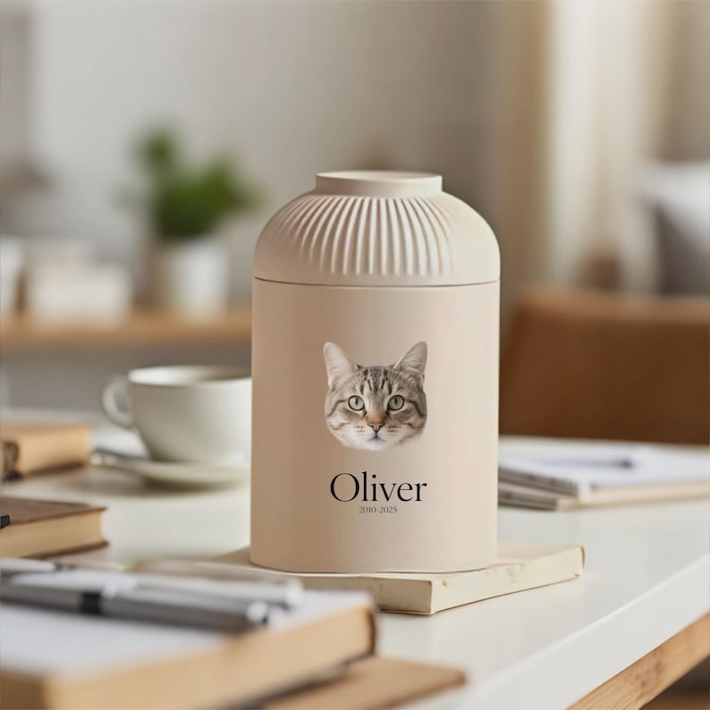 以下が含まれることがあります： 猫の顔と「Oliver」の名前がプリントされたベージュ色のペット用骨壺。骨壺はリブ付きのトップと円筒形をしています。名前の下のテキストは「2010-2025」です。