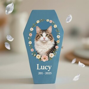 May include: A blue, coffin-shaped pet memorial box featuring a portrait of a cat surrounded by a floral wreath. The cat has brown and white fur. The name "Lucy" and the years "201-2025" are printed on the box.