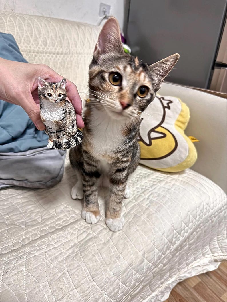 K&ouml;nnte beinhalten: Eine braune, wei&szlig;e und orange getigerte Katze sitzt auf einem wei&szlig;en Sofa. Eine kleinere, braune, wei&szlig;e und orange getigerte Katzenfigur wird in der Hand einer Person gehalten.
