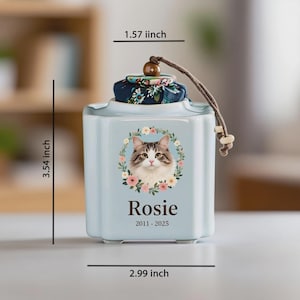 May include: A light blue pet urn with a floral wreath around a cat portrait. The urn is 3.54 inches tall, 2.99 inches wide, and 1.57 inches deep. The name "Rosie" and dates 2011-2025 are printed below the portrait. The lid has a floral fabric and a wooden bead.