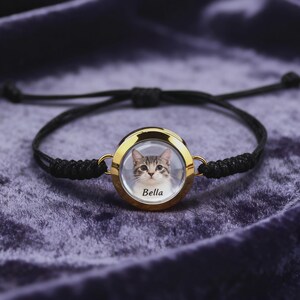 May include: A black adjustable bracelet with a gold-colored circular pendant. The pendant features a photo of a cat with the name "Bella" printed below the image. The bracelet is displayed on a purple velvet surface.