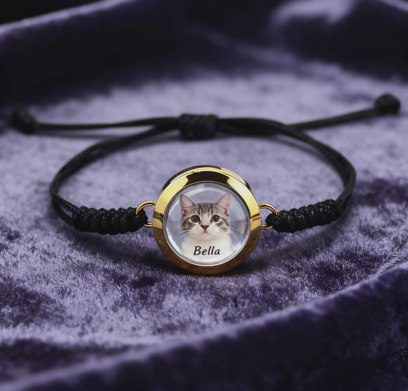 May include: A black adjustable bracelet with a gold-colored circular pendant. The pendant features a photo of a cat with the name "Bella" printed below the image. The bracelet is displayed on a purple velvet surface.