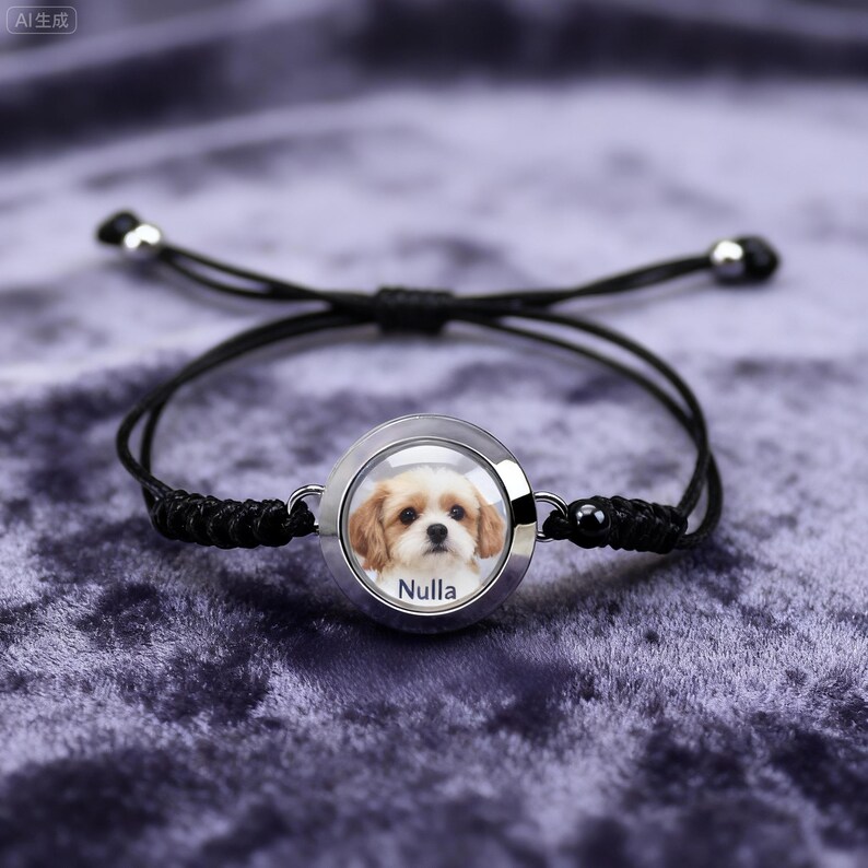 May include: Black adjustable bracelet featuring a circular pendant with a photo of a dog. The dog is light brown and white, with the name "Nulla" printed below. The bracelet has a woven cord design and silver-colored metal accents.