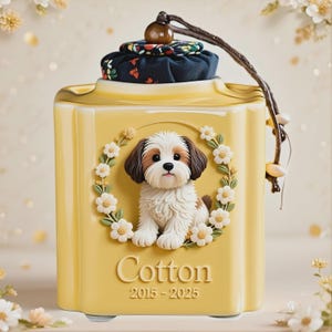 May include: Yellow ceramic container with a floral dog design. The container has a lid with a floral pattern and a brown cord. The word "Cotton" and the years "2015-2025" are printed on the front. The container is decorated with a wreath of white flowers.