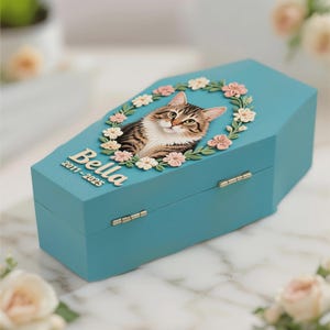 May include: A turquoise, coffin-shaped pet memorial box. The lid features a detailed relief of a tabby cat surrounded by a floral wreath. The name "Bella" and the years "2011-2025" are inscribed on the box.
