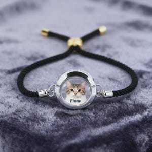 May include: A silver-toned bracelet with a black rope band. The focal point is a circular charm displaying a cat's portrait with the name "Finnn" below. The bracelet has gold-colored accents and a sliding knot closure.