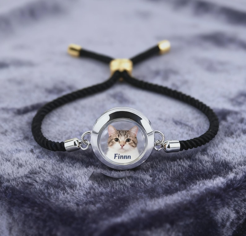 May include: A silver-toned bracelet with a black rope band. The focal point is a circular charm displaying a cat's portrait with the name "Finnn" below. The bracelet has gold-colored accents and a sliding knot closure.