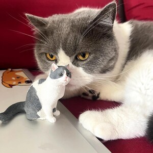 K&ouml;nnte beinhalten: Eine grau-wei&szlig;e Katze mit gelben Augen liegt auf einem roten Sofa. Eine kleine grau-wei&szlig;e Katzenfigur sitzt auf einem Laptop vor der Katze.