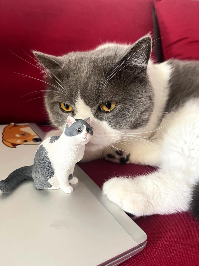 K&ouml;nnte beinhalten: Eine grau-wei&szlig;e Katze mit gelben Augen liegt auf einem roten Sofa. Eine kleine grau-wei&szlig;e Katzenfigur sitzt auf einem Laptop vor der Katze.