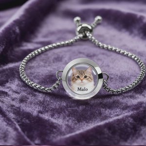 May include: Silver-toned bracelet with a round charm featuring a cat's portrait and the name "Malo." The charm is encased in clear glass. The bracelet is set against a purple velvet background.