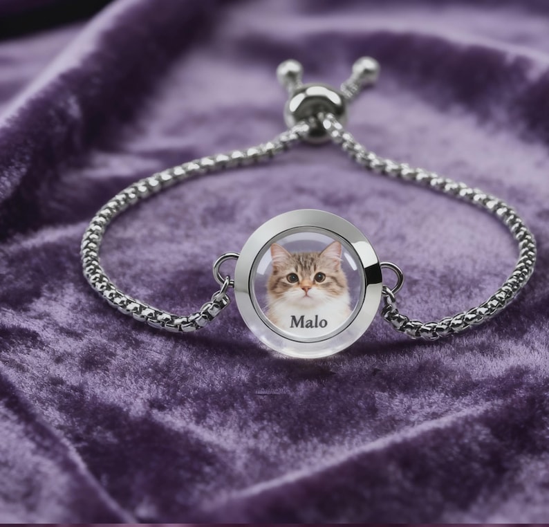 May include: Silver-toned bracelet with a round charm featuring a cat's portrait and the name "Malo." The charm is encased in clear glass. The bracelet is set against a purple velvet background.