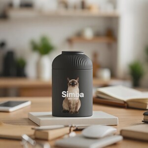 以下が含まれることがあります： サイアミーズ猫の画像と「Simba」の名前、白文字で「2013-2023」の年号が入った黒いペット用骨壺。 骨壺は円筒形で、リブ付きのトップがあり、白い正方形のベースの上に置かれています。