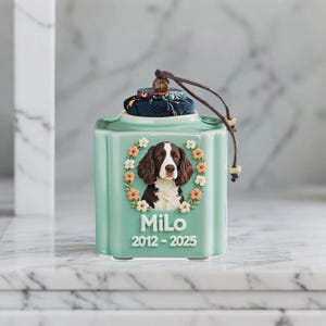 May include: A light green ceramic pet urn with a portrait of a dog surrounded by a floral wreath. The urn has the name "Milo" and the years "2012-2025" printed on it. The lid is topped with a patterned fabric and a brown cord.