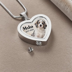 Pode incluir: Pingente prateado em forma de coração com corrente. O pingente apresenta uma foto colorida de um cachorro, o nome "Milo" e os anos "2011-2025". Um pequeno parafuso prateado está ao lado do pingente.