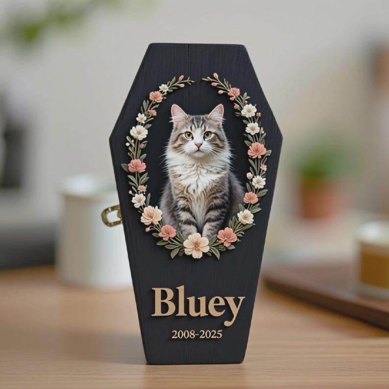 May include: A black wooden pet memorial in a coffin shape, featuring a portrait of a cat surrounded by a floral wreath. The cat is gray and white. The name "Bluey" and dates "2008-2025" are displayed in gold lettering.