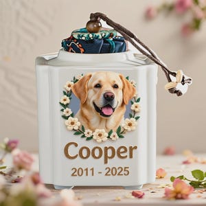 May include: A white ceramic pet urn featuring a portrait of a yellow Labrador Retriever surrounded by a floral wreath. The name "Cooper" and dates "2011-2025" are displayed below the portrait. The urn has a decorative lid with a patterned fabric.