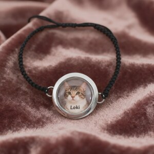May include: A black braided bracelet featuring a round, silver-toned pendant. The pendant contains a color photograph of a tabby cat with the name "Loki" printed below the cat's image. The bracelet is displayed on a pink velvet surface.