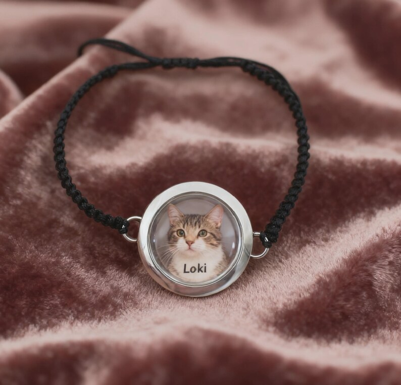May include: A black braided bracelet featuring a round, silver-toned pendant. The pendant contains a color photograph of a tabby cat with the name "Loki" printed below the cat's image. The bracelet is displayed on a pink velvet surface.