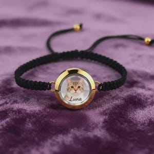 May include: A black braided bracelet with a gold-colored circular pendant. The pendant holds a photo of a cat with the name "Luna" printed below. The bracelet has adjustable black cords and gold-colored beads.