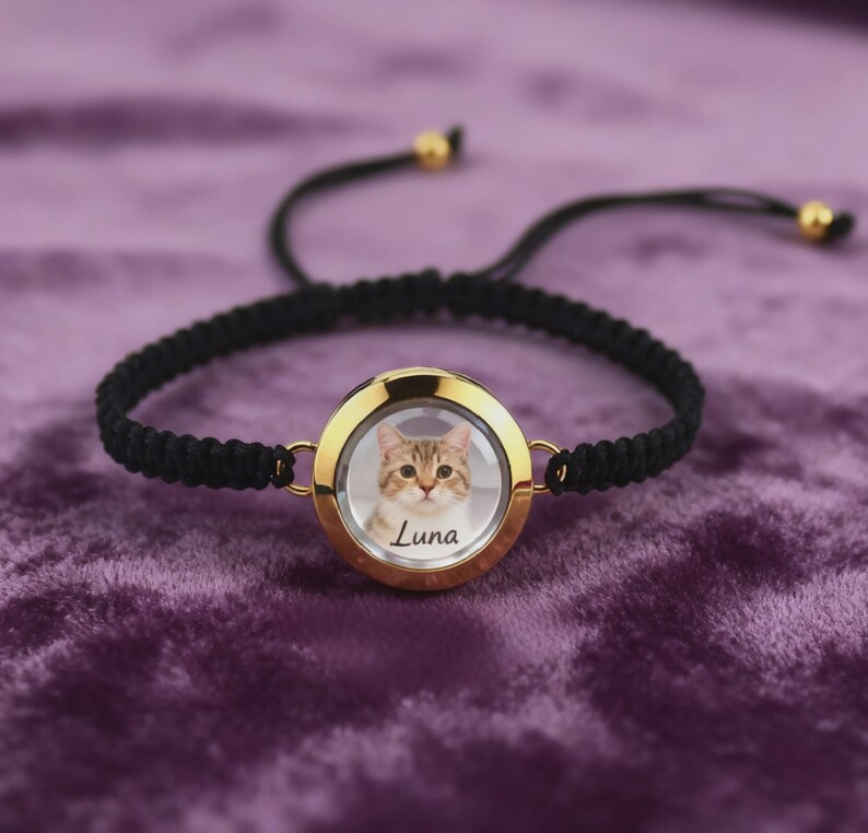 May include: A black braided bracelet with a gold-colored circular pendant. The pendant holds a photo of a cat with the name "Luna" printed below. The bracelet has adjustable black cords and gold-colored beads.