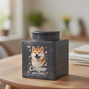 Peut inclure: Urne fun&eacute;raire carr&eacute;e noire avec un portrait de Shiba Inu, le nom "Cooper" et les ann&eacute;es 2012-2025. L'urne a un couvercle et des d&eacute;corations florales. L'urne est pos&eacute;e sur une table en bois.