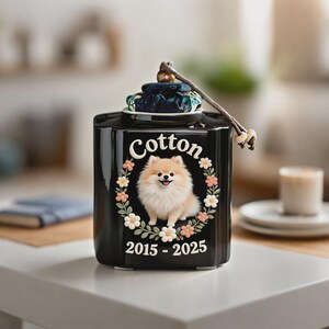 May include: Black ceramic pet urn with a Pomeranian illustration surrounded by a floral wreath. The word "Cotton" is above the dog, and the years "2015-2025" are below. The urn has a decorative lid with a fabric accent and a string handle.