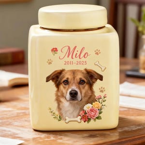 May include: A cream-coloured pet urn with a square shape and a matching lid. The urn features a portrait of a brown and white dog, the name "Milo", paw prints, and the years 2011-2025, with floral and bone accents.