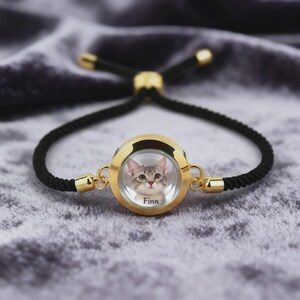 May include: A gold-toned bracelet with a black cord. The centerpiece is a round, gold-framed pendant containing a photo of a gray tabby cat named Finn. The bracelet has adjustable black cords and gold accents.
