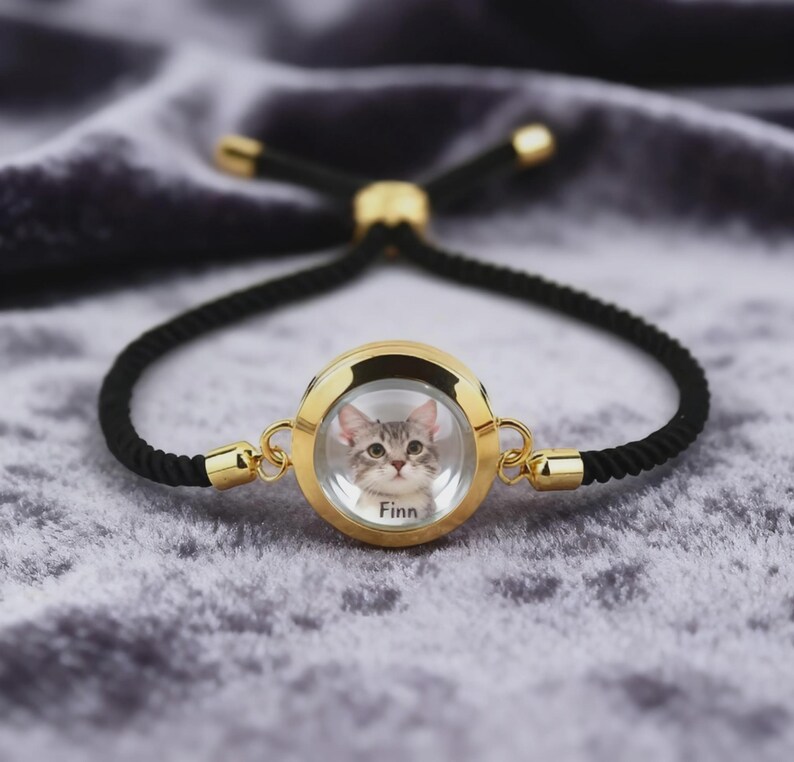May include: A gold-toned bracelet with a black cord. The centerpiece is a round, gold-framed pendant containing a photo of a gray tabby cat named Finn. The bracelet has adjustable black cords and gold accents.