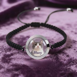 May include: Black braided bracelet with a circular, silver-toned pendant. The pendant features a photo of a gray and white cat with the name "Bluey" printed below. The bracelet has adjustable black cords.