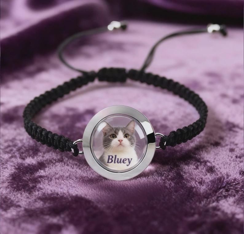 May include: Black braided bracelet with a circular, silver-toned pendant. The pendant features a photo of a gray and white cat with the name "Bluey" printed below. The bracelet has adjustable black cords.