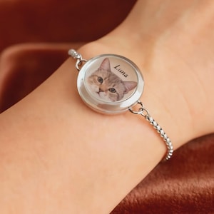 May include: Silver bracelet featuring a round charm with a cat portrait and the name "Luna". The cat has brown and tan fur and green eyes. The bracelet is worn on a wrist.
