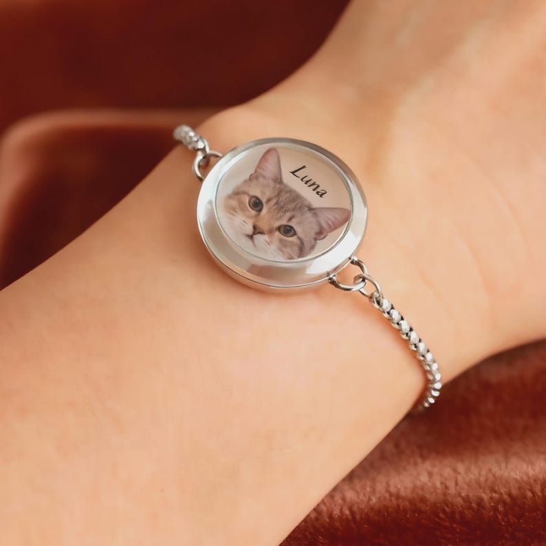 May include: Silver bracelet featuring a round charm with a cat portrait and the name "Luna". The cat has brown and tan fur and green eyes. The bracelet is worn on a wrist.