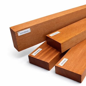 May include: Four rectangular mahogany wood planks with a reddish-brown hue are displayed. Each plank has a smooth, polished surface. Small white labels with black text are affixed to the ends of the planks. The planks are stacked and arranged to show their different sizes.