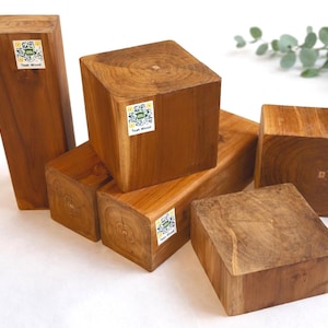 May include: A collection of teak wood blocks in various sizes and shapes. The blocks are a warm brown color, with visible wood grain and some with stickers that read "Teak Wood". The blocks are arranged on a white surface.