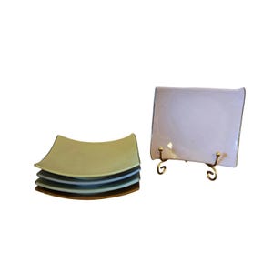 May include: A stack of four square ceramic plates in shades of light green, blue, teal, and brown, positioned next to a single light purple square plate displayed upright on a gold-toned metal wire stand with curved decorative feet.