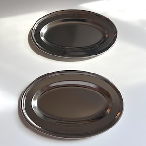 May include: Two oval-shaped, stainless steel serving platters. The platters have a polished, reflective surface and a slightly raised rim. They are placed on a white surface, with natural light casting shadows.
